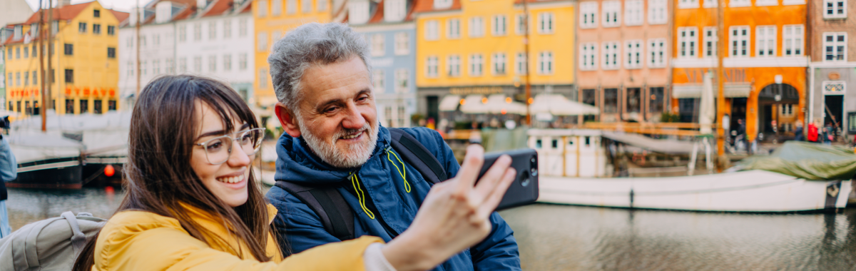woman on a trip to Copenhagen with a taking a selfie with her friend with a harbor full of sailboats 