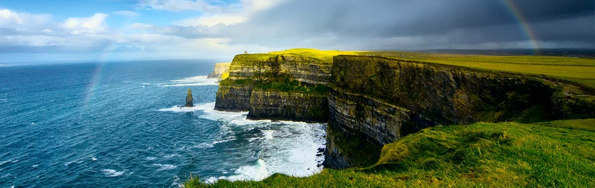 Scenic Irish vista with green grass, a rainbow, cliffs and ocean that makes you want to travel to Ireland